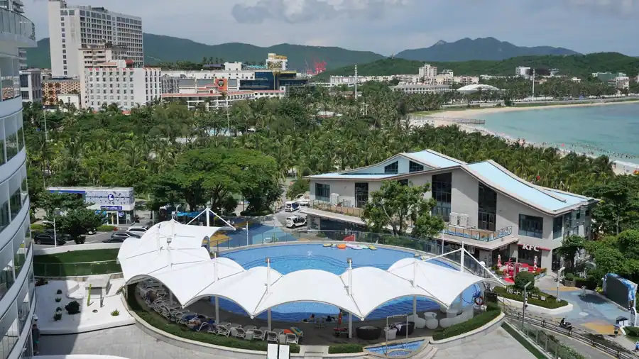Aerial view of Sanya's beachfront with pool, buildings, and lush greenery under a clear sky.